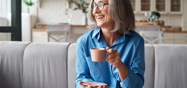 Happy relaxed mature older adult grey-haired woman drinking coffee relaxing on sofa at home